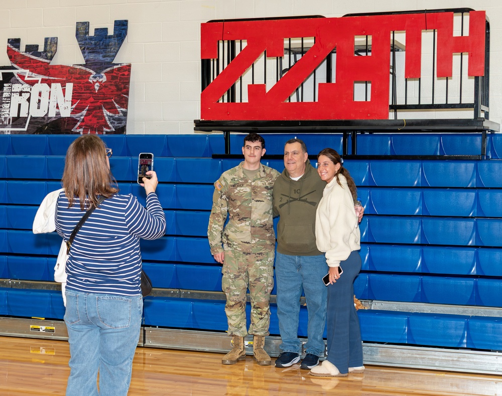 A woman in a striped shirt photographs three people posing in front of blue gymnasium bleachers. The group includes a young person in U.S. Army camouflage uniform flanked by two civilians, suggesting a military family reunion or send-off event. The setting appears to be a school or community gymnasium decorated with team or unit insignia.