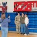 A woman in a striped shirt photographs three people posing in front of blue gymnasium bleachers. The group includes a young person in U.S. Army camouflage uniform flanked by two civilians, suggesting a military family reunion or send-off event. The setting appears to be a school or community gymnasium decorated with team or unit insignia.