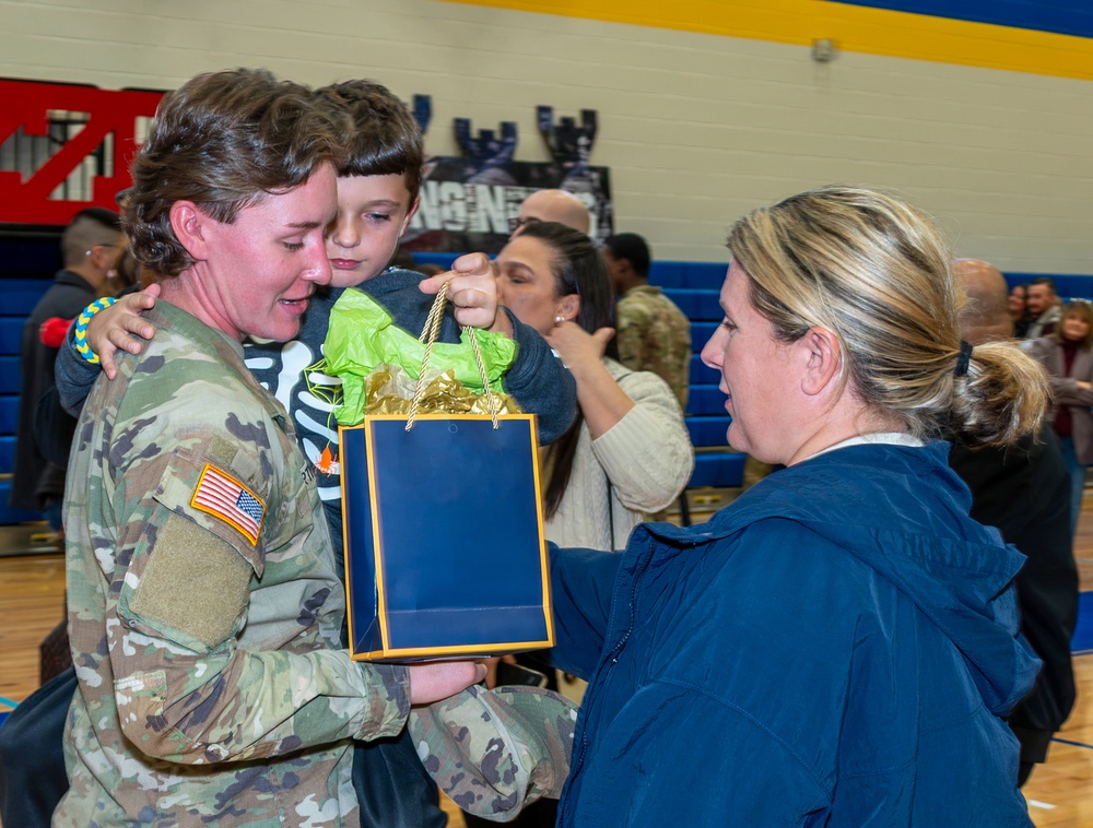 A female U.S. Army soldier in OCP camouflage uniform is being reunited with a young child who is embracing her, while a civilian woman presents her with a gift bag. The event appears to be a military homecoming or welcome ceremony taking place inside a school or community gymnasium. The emotional scene captures the joy of a military reunion with family members and other personnel visible in the background.