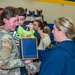 A female U.S. Army soldier in OCP camouflage uniform is being reunited with a young child who is embracing her, while a civilian woman presents her with a gift bag. The event appears to be a military homecoming or welcome ceremony taking place inside a school or community gymnasium. The emotional scene captures the joy of a military reunion with family members and other personnel visible in the background.