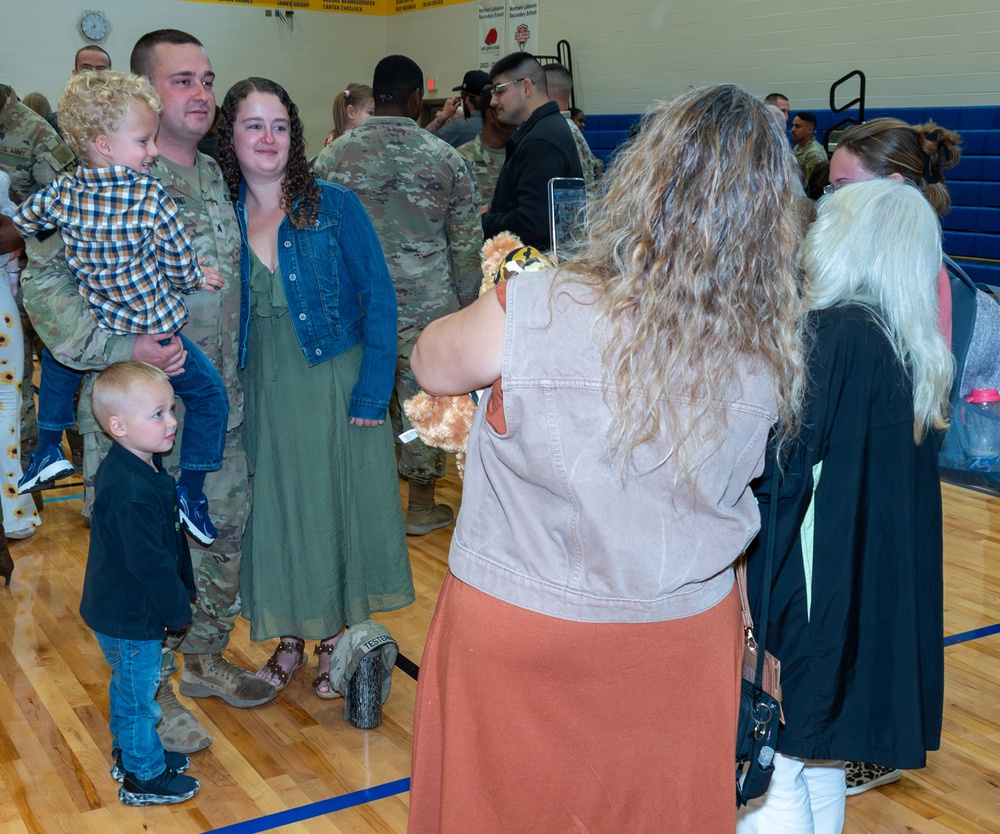 A military homecoming or deployment ceremony taking place inside a school gymnasium. A U.S. Army soldier in camouflage uniform is reuniting with his family, holding a young child while standing next to a woman and another small child. A person in the foreground is photographing the family reunion moment.