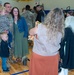 A military homecoming or deployment ceremony taking place inside a school gymnasium. A U.S. Army soldier in camouflage uniform is reuniting with his family, holding a young child while standing next to a woman and another small child. A person in the foreground is photographing the family reunion moment.