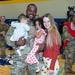 A U.S. Army soldier in OCP camouflage uniform is reuniting with his family in what appears to be a military gymnasium or community center. He is holding two young children — an infant and a toddler — while standing beside a smiling woman, likely his spouse. The setting suggests a military homecoming or welcome-back ceremony.