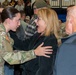A female U.S. Army soldier in OCP (Operational Camouflage Pattern) uniform is greeting a civilian woman who appears overjoyed and surprised, likely at a military homecoming or deployment ceremony held in a gymnasium. The civilian woman's expression of shock and happiness, combined with the soldier's hand on her chest, suggests an emotional reunion between family members or close friends. The background shows a crowded indoor venue with other soldiers and civilians present.