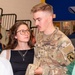 A U.S. Army soldier in OCP (Operational Camouflage Pattern) uniform is engaged in conversation with a young woman wearing glasses and a black top with layered necklaces. The soldier's uniform displays 'U.S. ARMY' tape, a name tape partially reading 'SH-U', a Private First Class rank insignia, and a shoulder patch featuring a horse (likely the 1st Cavalry Division). The setting appears to be an indoor gymnasium or community hall with bleachers visible in the background.