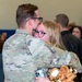 Two individuals in military camouflage uniforms are embracing in what appears to be an indoor facility, likely during a military homecoming or deployment ceremony. The person facing the camera is a young woman with blonde hair and glasses, visibly emotional with red eyes, holding a stuffed teddy bear dressed in camouflage clothing. Additional people are visible in the blurred background.