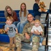 A U.S. Army soldier named Raber, wearing OCP (Operational Camouflage Pattern) uniform, is seated on gymnasium bleachers surrounded by family members. He is holding a young toddler on his lap while a young boy stands beside him, with two women and an older child seated behind them. The setting appears to be a military homecoming or family event held in a school or community gymnasium.