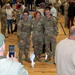 A military gathering or homecoming event taking place inside a gymnasium, where U.S. Army soldiers in OCP (Operational Camouflage Pattern) uniforms are reuniting with family members and civilians. Three soldiers are posing for a photo in the foreground while a civilian photographs them with a smartphone. The scene shows widespread emotional reunions and group interactions throughout the gym.