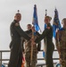 A military change of command ceremony is taking place, with two officers in olive green flight suits exchanging a unit guidon (flag). Several personnel in OCP (Operational Camouflage Pattern) uniforms stand in the background alongside blue Air Force unit flags. The visible flag in the background partially reads '274 AW', suggesting this is an Air Wing unit ceremony.