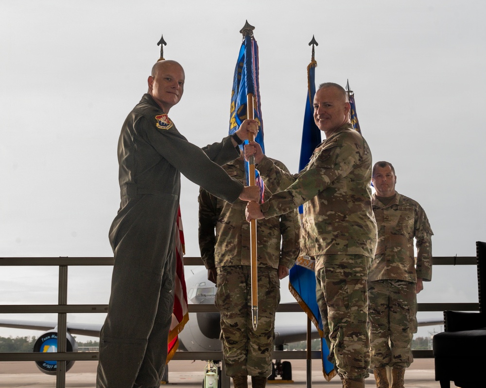 A military change of command ceremony is taking place on an airfield tarmac, with two senior officers exchanging a guidon (unit flag on a staff). The officer on the left is wearing an olive green flight suit, while the officer on the right is in OCP (Operational Camouflage Pattern) uniform. Additional military personnel stand in formation in the background near blue unit flags and what appears to be a large military aircraft.