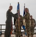 A military change of command ceremony is taking place on an airfield tarmac, with two senior officers exchanging a guidon (unit flag on a staff). The officer on the left is wearing an olive green flight suit, while the officer on the right is in OCP (Operational Camouflage Pattern) uniform. Additional military personnel stand in formation in the background near blue unit flags and what appears to be a large military aircraft.