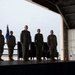 A formal military ceremony is taking place inside an aircraft hangar, with five uniformed personnel standing at attention on a raised stage or platform. Two individuals are wearing olive green flight suits while others are in standard camouflage uniforms. A blue Air Force flag with gold fringe is prominently displayed on the left side of the stage.