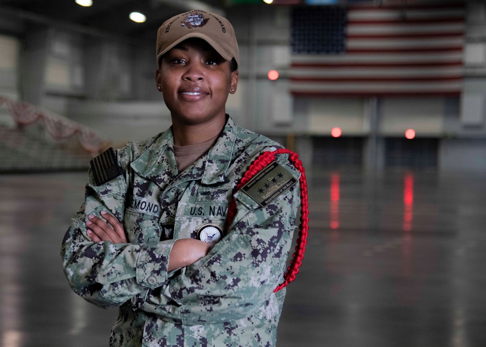 A U.S. Navy service member poses with arms crossed in what appears to be a large hangar bay, likely aboard a naval vessel or naval installation. The individual is wearing the Navy Working Uniform (NWU) with visible insignia including a name tape reading partial 'EMOND' and 'U.S. NAVY' tape, along with a red aiguillette and various patches. An American flag is visible in the background.