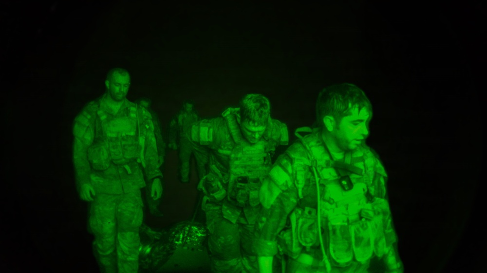 A night vision image showing multiple military personnel in full tactical gear moving through darkness. The green monochromatic imagery is characteristic of night vision optics or photography through night vision equipment. At least four individuals are visible, wearing body armor and carrying equipment.