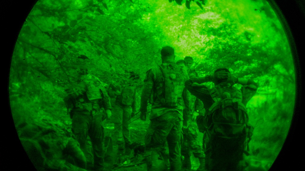 The image is captured through a night vision device, showing a circular green-tinted field of view. Multiple individuals in military-style gear and camouflage uniforms are visible in what appears to be a wooded or forested environment. The scene suggests a nighttime military patrol, training exercise, or tactical operation.