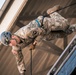 A U.S. Army soldier is performing a rappelling exercise off the edge of a building or training tower. The soldier is wearing full military combat uniform with visible unit patches and is secured by a rope and harness system. This appears to be a military training exercise focused on vertical descent techniques.