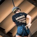 A person is rappelling or preparing to descend from a structure, photographed from below looking upward. They are wearing safety gear including a helmet and harness, along with a black 'Band of Brothers' t-shirt and blue shorts. The upward angle provides a unique perspective of the climber against what appears to be the underside of a metal or industrial structure.