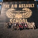 A large group of approximately 30-40 individuals poses in front of a large wooden building bearing the text 'THE AIR ASSAULT SCHOOL' with the Air Assault Badge insignia painted in gold on a black background. The group includes a mix of military personnel in camouflage uniforms and civilians in casual clothing. The photo appears to be an official or commemorative group photograph taken at a U.S. Army Air Assault School facility.