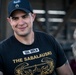 A smiling male individual wearing a black baseball cap with a military patch and a black t-shirt with the text 'SSG BERLIN' and 'THE SABALAUSKI' along with a gold Combat Infantryman Badge (CIB) graphic. The cap displays what appears to be a Staff Sergeant rank insignia patch. A second person in camouflage uniform is visible but blurred in the background.