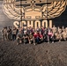 A large group of approximately 30 people poses for a group photo in front of a dark wooden wall featuring a large mural with wings, a crown, and the word 'SCHOOL'. The group includes both military personnel in camouflage uniforms and civilians in casual attire. They are arranged in two rows — standing in the back and kneeling in the front — on a gravel surface.