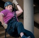 A middle-aged man is rappelling or abseiling down a rope near a large structural column, likely on the exterior of a building. He is wearing full safety gear including a helmet, harness, and gloves. The image is taken from a low angle, emphasizing the height and physical effort involved in the activity.