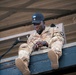A U.S. Army soldier wearing multicam camouflage uniform sits on the edge of what appears to be a raised platform or structure. The soldier, identified by name tape as 'FOX,' is wearing an Airborne division patch on the right shoulder and a black baseball cap. A black rope is visible nearby, suggesting a rappelling or obstacle course activity.