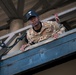 A U.S. Army soldier wearing multicam/OCP camouflage uniform is photographed from a low angle as he leans over a metal railing or platform edge. His name tape reads 'COLEMAN' and he wears a Sergeant rank insignia along with a black baseball cap bearing a military insignia. The image is taken from below, giving a dramatic upward perspective of the soldier and the structure above him.