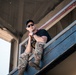 A man is seated on a metal railing or ledge of an elevated structure, photographed from a low angle looking upward. He is wearing casual-tactical clothing and appears relaxed in a posed or candid moment. The industrial or stadium-like structure behind him provides a gritty urban backdrop.