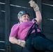 A woman is rappelling or zip-lining while suspended by a harness and rope system against what appears to be a large metal structure. She is smiling and appears confident, gripping the rope with both gloved hands. She is equipped with proper safety gear including a helmet, harness, and gloves.
