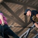 A person in a pink shirt and helmet is rappelling or preparing to descend from an elevated structure while an instructor in a black cap and t-shirt supervises from the edge. The setting appears to be a military or adventure training facility, suggested by the instructor's shirt referencing 'Air Assault School' and the participant's shirt reading 'Band of Brothers, Fort Campbell, KY'. Safety equipment including a harness, rope, gloves, and carabiner hardware are clearly visible.