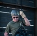 A middle-aged man is engaged in rappelling or rope descent activity, wearing full safety gear including a helmet and harness. He is gripping a dark rope with a gloved hand while a carabiner is visible attached to his harness. The industrial metal structure in the background suggests an urban or warehouse rappelling environment.