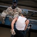 A U.S. Army soldier in OCP (Operational Camouflage Pattern) uniform, identified by name tape as 'FOX' with Staff Sergeant rank insignia, sits at the edge of an elevated platform supervising a civilian participant in a rappelling activity. The civilian, wearing a white sleeveless shirt, black leggings, a safety helmet, rappelling harness, and leather gloves, is descending a rope from the structure. This appears to be a military outreach or public engagement event where civilians experience rappelling under military supervision.
