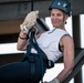 An older woman is engaged in rappelling or abseiling activity, suspended by a rope and harness system. She is wearing professional climbing gear including a Black Diamond helmet, work gloves, and a full body harness with carabiners. The setting appears to be an urban or industrial structure based on the steel beams visible in the background.