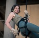 A woman is rappelling or zip-lining outdoors, suspended in a full-body harness attached to a rope. She is smiling broadly with her eyes closed, suggesting enjoyment or excitement during the activity. The setting appears to be near a concrete structure, possibly a bridge or parking garage.