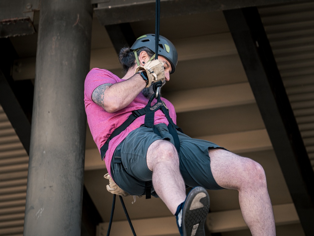 A man is suspended mid-air on a rope system, appearing to be rappelling or zip-lining beneath a large industrial or recreational structure. He is wearing safety equipment including a helmet and harness, and is actively gripping the rope with gloved hands. The photo is taken from a low angle looking upward, emphasizing his suspended position.