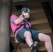 A man is suspended mid-air on a rope system, appearing to be rappelling or zip-lining beneath a large industrial or recreational structure. He is wearing safety equipment including a helmet and harness, and is actively gripping the rope with gloved hands. The photo is taken from a low angle looking upward, emphasizing his suspended position.