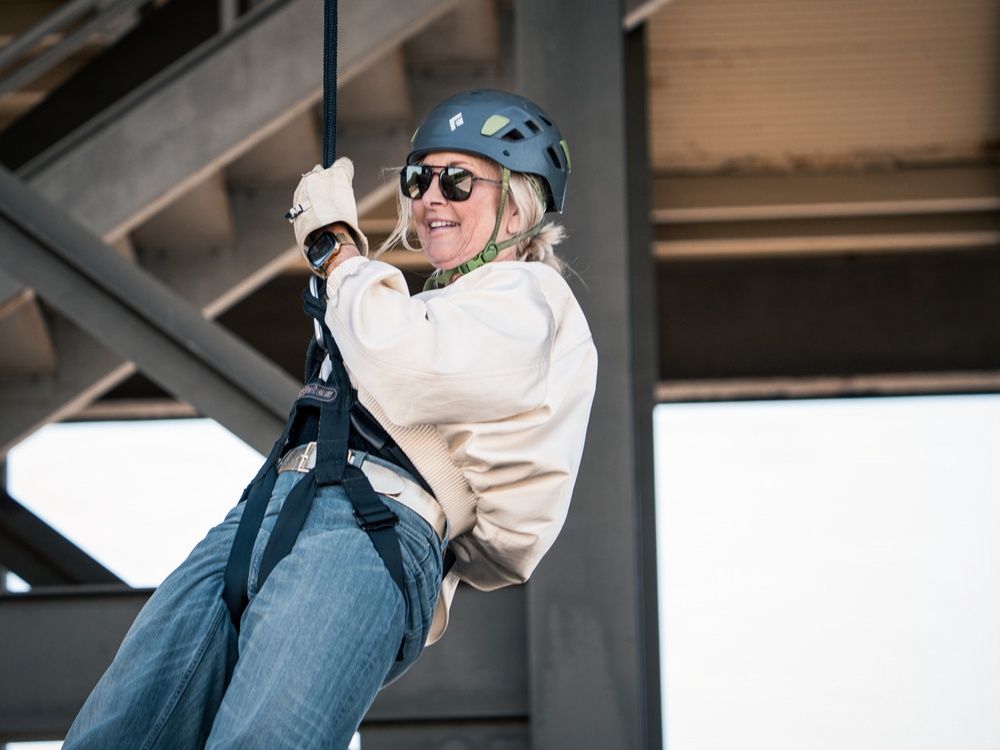 A woman is suspended on a rope or zip line beneath a large steel structure, wearing full safety equipment including a helmet and harness. She appears to be smiling and enjoying the activity, gripping the rope with gloved hands. The setting appears to be an urban or industrial environment based on the visible steel framework overhead.