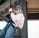 A woman is suspended on a rope or zip line beneath a large steel structure, wearing full safety equipment including a helmet and harness. She appears to be smiling and enjoying the activity, gripping the rope with gloved hands. The setting appears to be an urban or industrial environment based on the visible steel framework overhead.