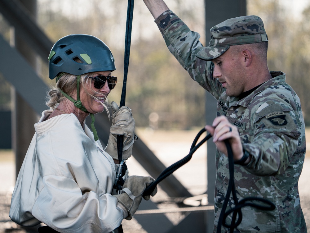 A civilian woman wearing a blue climbing helmet, sunglasses, and work gloves is being assisted by a U.S. Army soldier in full multicam uniform during what appears to be a rappelling or rope descent activity. The soldier, who has an Airborne patch visible on his uniform, is managing the rope and providing instruction or guidance. The setting appears to be a military training facility or obstacle course.