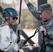 A civilian woman wearing a blue climbing helmet, sunglasses, and work gloves is being assisted by a U.S. Army soldier in full multicam uniform during what appears to be a rappelling or rope descent activity. The soldier, who has an Airborne patch visible on his uniform, is managing the rope and providing instruction or guidance. The setting appears to be a military training facility or obstacle course.