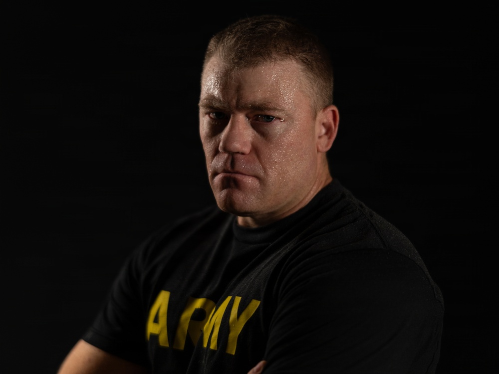 A muscular male individual is photographed against a dark black background in a dramatic, low-key portrait style. He is wearing a black t-shirt with 'ARMY' printed in yellow letters and has his arms crossed. His expression is intense and serious, and his skin appears to have a slight sheen, suggesting recent physical exertion.