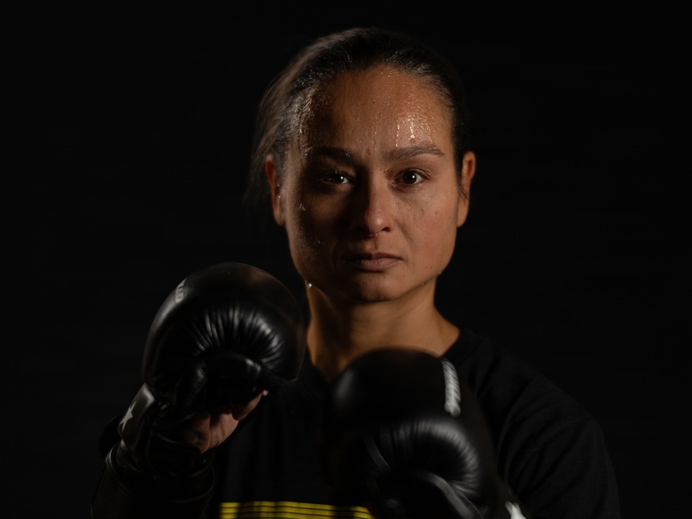 A female boxer is photographed in a dramatic, low-key studio portrait against a black background. She is wearing black boxing gloves and a black athletic top, with her fists raised in a guard position. Her face is visibly covered in sweat, suggesting recent intense physical exertion, and her expression is focused and determined.