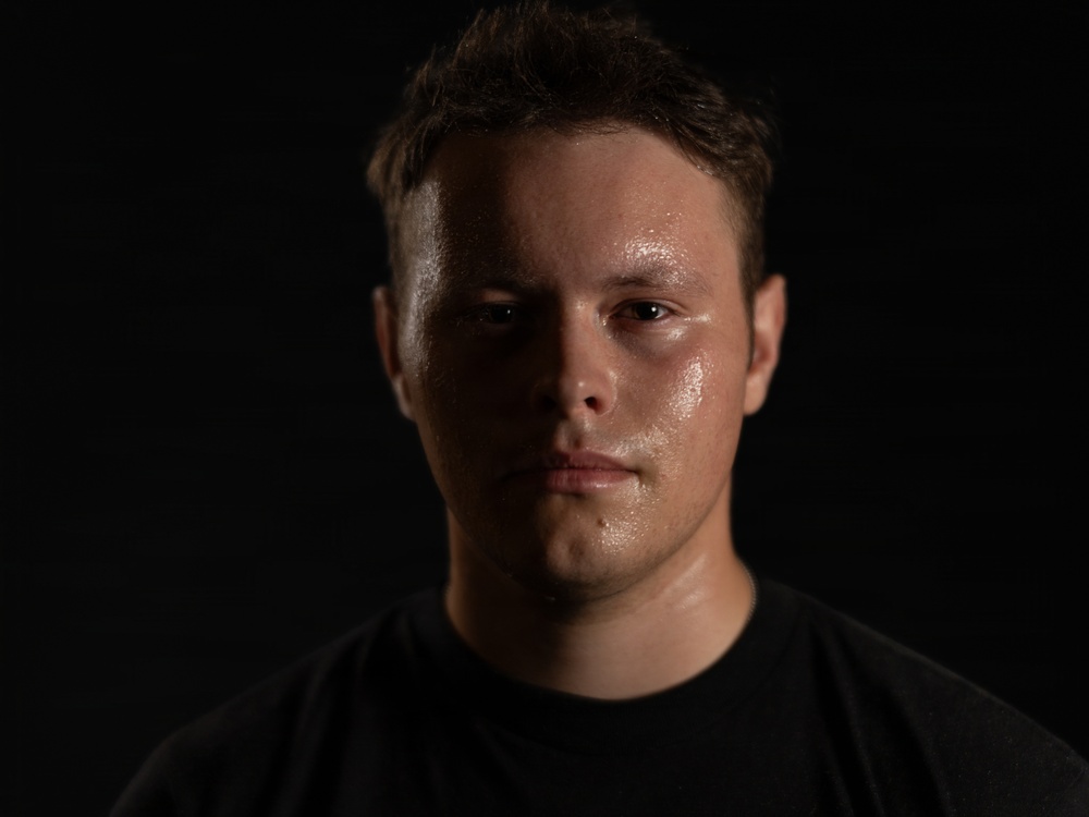 A close-up portrait of a young adult male against a completely black background, lit with dramatic directional lighting. His face appears visibly sweaty or moist, with light reflecting off his skin. His expression is neutral to serious, and he is wearing a black t-shirt.