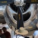 The image shows visitors at an airshow examining the engine and propeller assembly of a vintage aircraft up close. Yellow stenciled markings on the propeller blade are legible, identifying blade specifications including 'BLADE DWG. GS21A-G', 'BLADE MFG. FCG24', 'LOW ANGLE 19.0 AT 72', and 'HIGH ANGLE 84.0 AT 72'. The polished chrome engine cowling reflects the surrounding environment, and multiple spectators are gathered around the aircraft.