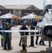 An outdoor airshow event at what appears to be Vance Air Force Base, featuring a highly polished chrome aircraft with 'VANCE' written on its tail section. Crowds of visitors are gathered around the static display aircraft on the tarmac, with vendor tents and hangars visible in the background. American flags and commercial banners including 'Budweiser' are visible among the event infrastructure.