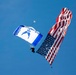 A U.S. Air Force parachutist is descending under a blue and white canopy bearing the U.S. Air Force logo and text. The skydiver is carrying a large American flag that billows dramatically in the wind against a clear blue sky. This appears to be a ceremonial or airshow demonstration jump.