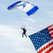 A U.S. Air Force parachutist descends under a blue and white canopy marked with 'U.S. Air Force' and 'airforce.com' branding. The jumper is carrying a large American flag, which billows dramatically in the foreground. This appears to be a ceremonial or airshow demonstration jump.