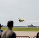 An airshow is taking place at an airfield where a yellow biplane performs a low-altitude aerobatic maneuver, trailing white smoke. Spectators stand behind metal crowd-control barriers in the foreground watching the performance. Airport infrastructure including hangars and a radar dome are visible in the background.