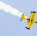 A bright yellow aerobatic biplane is captured mid-maneuver against a clear blue sky, trailing a white smoke stream behind it. The aircraft is inverted or in a steep bank, showcasing an airshow performance. The name 'Brian Correll' is visible on the fuselage, identifying the pilot or aircraft owner.