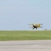 A yellow and black biplane is flying at very low altitude between two poles connected by a rope or ribbon with red flags at each end, suggesting a ribbon-cutting or low-level precision flying demonstration at an airshow. Ground crew members are stationed at each pole on either side of the aircraft. The scene takes place on a grass field adjacent to a paved runway under a clear sky.